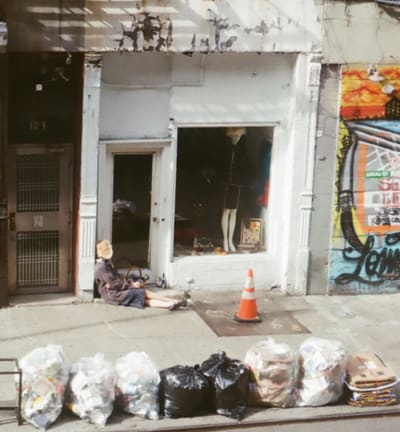 Storefront with a mannequin, a person seated on the sidewalk, an orange cone and a row of trash bags
