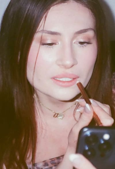 Film photo of a person with long brown hair applying lip liner while looking down at a phone