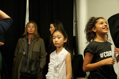Two young models and adults standing in a room with black curtains