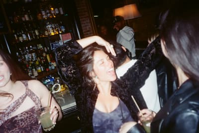 Laughing person in a black velvet jacket with an arm over their head at a busy bar