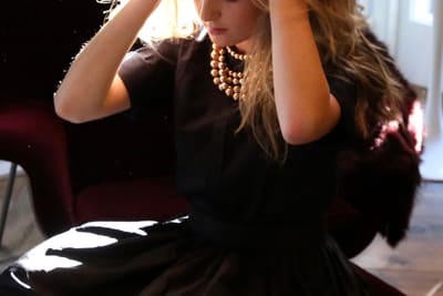 Backlit model in a black dress and pearl necklace with hands raised to their hair