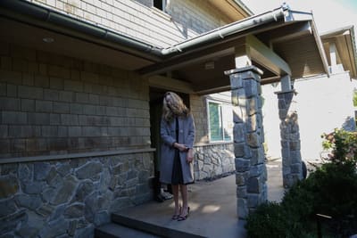 Model with long blonde hair in a grey coat standing on the stone porch of a house