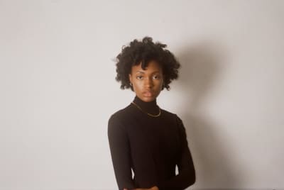 Person in a black turtleneck and gold chain, arms folded, against a plain pale wall