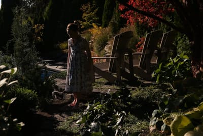 Young model in a floral dress walking through a shaded garden past wooden deck chairs