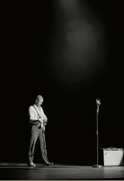 Black-and-white photo of a lone performer with a guitar on a dark stage under a single spotlight